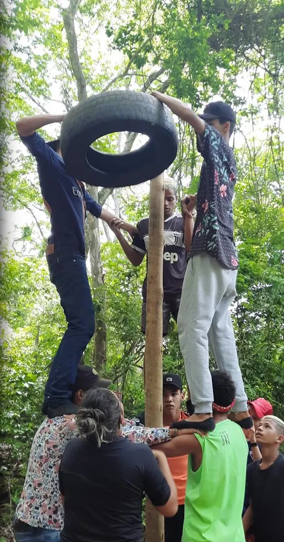 Een groep jongeren tilt samen een autoband over een houten paal, terwijl anderen hen ondersteunen.